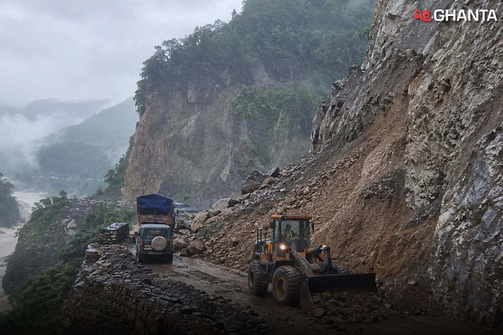 Heavy machinery clears landslide debris from a narrow, rain-soaked road as traffic waits to pass.